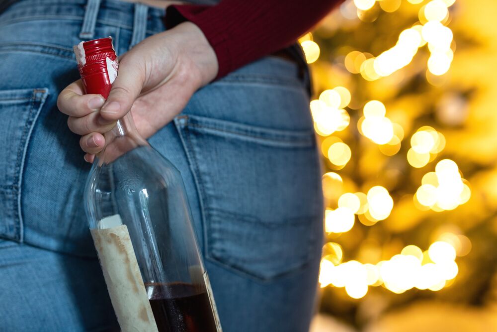 Woman concealing a wine bottle behind her near a Christmas tree with festive lights, representing secretive drinking tied to holiday stress.