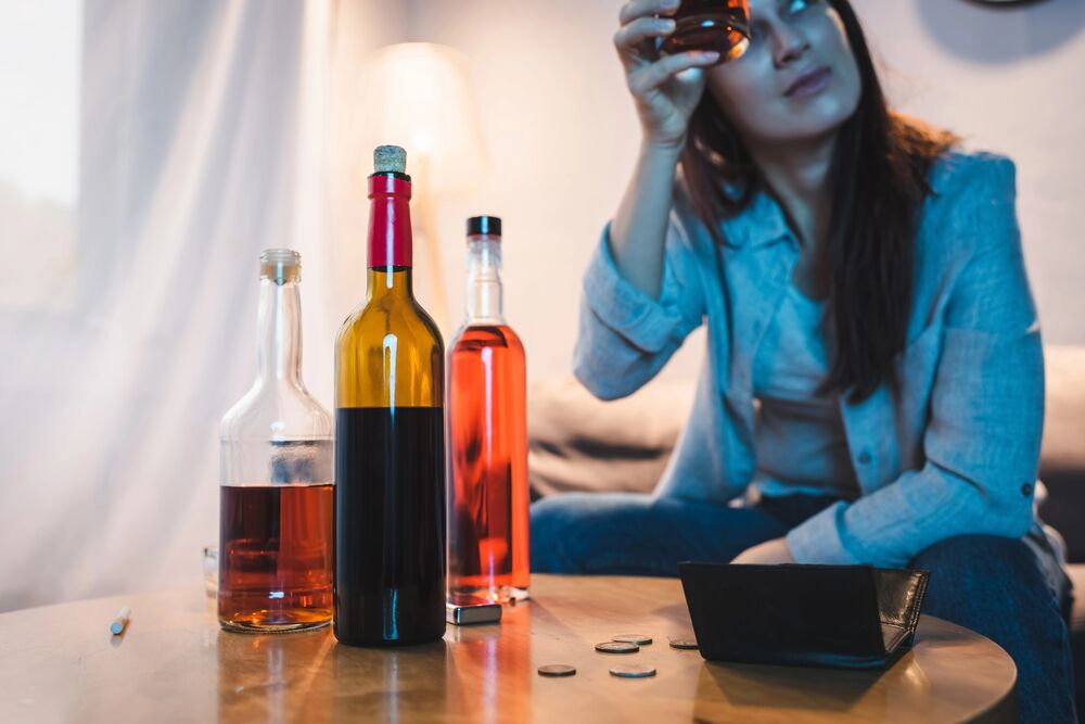 Image Alt: woman drinking alcohol while surrounded by bottles and loose change, symbolizing how rising expenses can contribute to unhealthy coping behaviors.